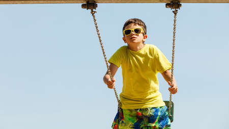 Rest and relax for children. Little boy in sunglasses resting swinging outdoor. Adorable child having fun playing in playground.の写真素材