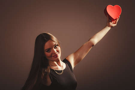Love help and charity concept. Young beauty cute happy long haired woman girl in black dress holding heart box gift present in studio.の写真素材