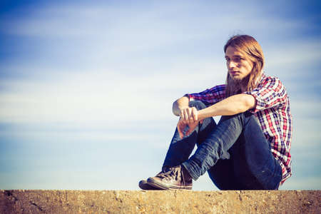 Man long hair wearing plaid shirt relaxing outdoor sitting on concrete wall at sunny windy day against blue skyの写真素材