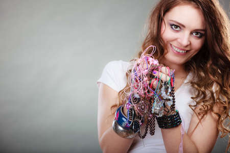 Pretty young woman wearing bracelets and rings holding many plentiful of precious jewelry necklaces beads. Portrait of gorgeous fashion girl in studio on gray.の写真素材