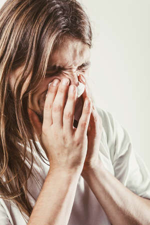 Allergy and cold concept. Young long haired sick man with hygienic tissue. Guy blowing nose.の写真素材
