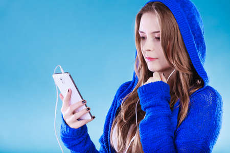 young woman with smart phone listening music. Teen stylish long hair girl in hood relaxing or learning language. Studio shot on blue.の写真素材