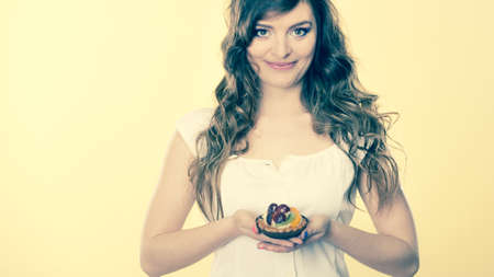 Bakery, sweet food and people concept. smiling cute woman curly hair holding fruit cake cupcake in hand yellow backgroundの写真素材