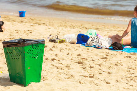 Order and cleanliness at the beach. Tourists resting lying on seaside. Green metal trash can on sand.の写真素材