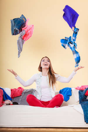 Happy woman sitting on sofa couch in messy living room throwing clothes. Young girl surrounded by many stack of clothing. Disorder and mess at home.の写真素材