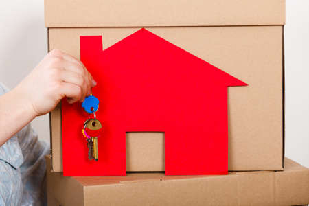 Closeup of human hand holding keys to new apartment. Red paper house and boxes in background. Real estate and housing concept.の写真素材