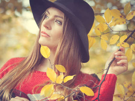 Autumn season colors in the park concept. Woman standing by the tree. Attractive lady wearing black hat and red sweather and very stylish checkered scarf.の写真素材