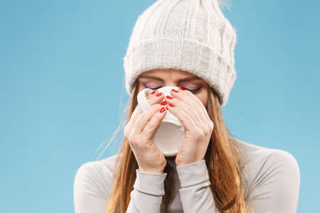 Attractive woman long hair girl in winter wool cap holds mug with tea hot beverage, warming herself studio shot on blue.の写真素材