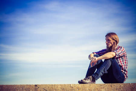 Man long hair wearing plaid shirt relaxing outdoor sitting on concrete wall at sunny windy day against blue skyの写真素材