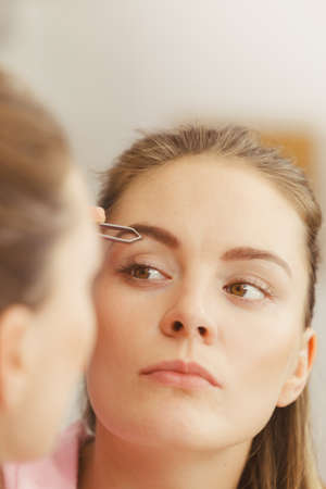 Woman plucking eyebrows depilating with tweezers. Attractive girl tweezing eye brows looking in bathroom mirror.の写真素材