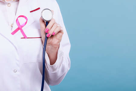 Women fight for health. Breast cancer tumor concept. Pink ribbon and blue stethoscope on white medical apron uniform. Doctor holding medic equipment.の写真素材