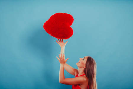 Pretty cute young woman girl throwing red heart shape pillow in studio on blue. Valentines day love.の写真素材