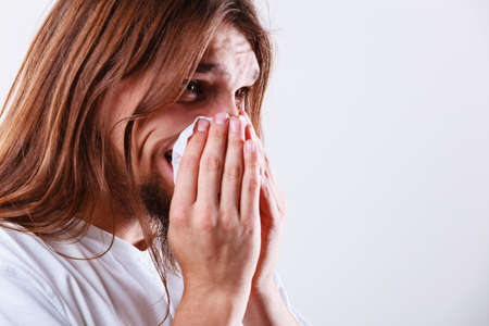 Allergy and cold concept. Young long haired sick man with hygienic tissue. Guy blowing nose.