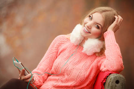 Digital equipment, student outside. Young woman with cell phone. Attractive girl sitting on the bench in park. Autumnal weather.の写真素材