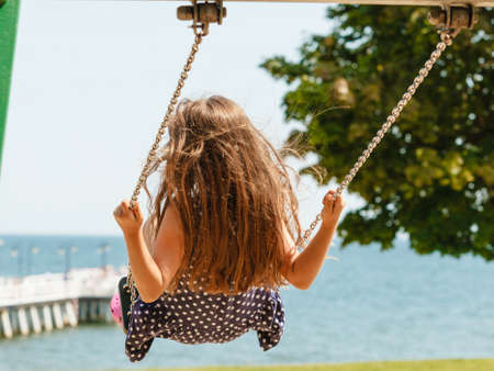 Have fun and leisure concept. Long haired enjoyable girl swinging outdoor in garden playground. Lovely child playing on swing-set.の写真素材