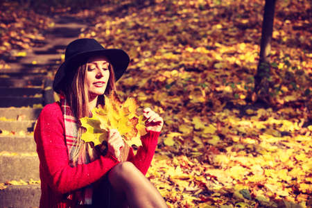 Woman relaxing in autumn fall park. Young feshionable female stylish black hat holding yellow maple leaves in hand, enjoying sunlightの写真素材