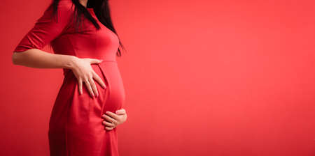 New life concept. Pregnancy, motherhood and happiness. Closeup on tummy of pregnant woman wearing stylish elegant red dress indoorの写真素材