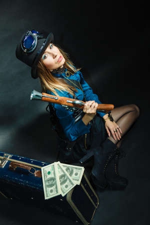 Fashionable subculture weapon concept. Steampunk girl with cash. Young gorgeous lady in victorian fashion lying on floor with banknotes briefcase and blunderbuss.の写真素材