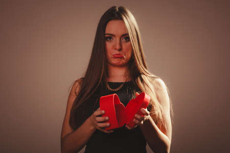 Valentine day. Sad upset grymacing woman in black opening empty heart box gift present. Elegant attractive model with valentines symbol sign. Studio shot.の写真素材