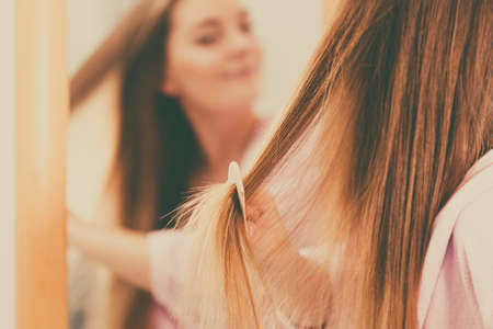 Woman combing brushing her long smooth hair in bathroom, looking in mirror. Girl taking care refreshing her hairstyle. Haircare concept.の写真素材