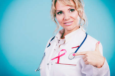 Breast cancer. Medical help treatment concept. Female blonde doctor in white medic apron showing pink ribbon and stethoscope on blue background.の写真素材