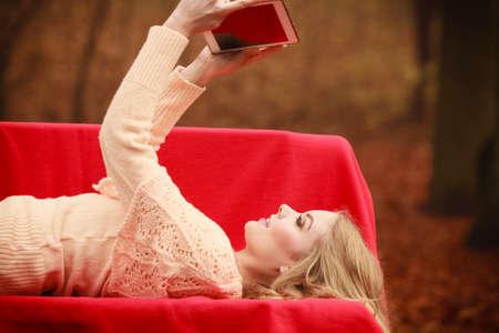 Young blonde girl in park. Cheerful lady sitting on bench browsing her tablet surrounded by autumnal vegetation.の写真素材