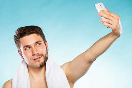 Young man with half shaved face beard hair taking selfie self photo with smartphone camera. Handsome guy on blue. Skin care and hygiene.の写真素材