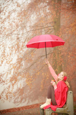 Nature outdoor weather fun cozy concept. Girl sitting in park. Lady sovering herself from rain by holding umbrella drinking coffee from cup while staying on bench.の写真素材