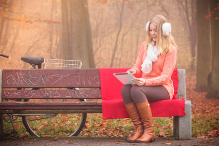 Autumn time people outside. Beautiful woman with digital tablet. Young lady sitting on the bench and wearing autumnal clothing.の写真素材