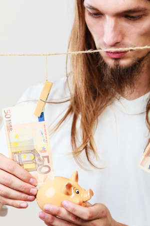Savings concept. Young handsome long haired man holding piggybank moneybox around banknotes cash.