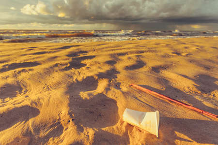 Polluted beach shore. Plastic disposable cup abandoned on sand, beautiful golden sunset in the background. Earth ecologyの写真素材