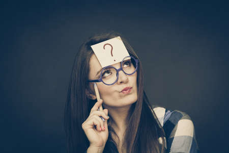Woman confused thinking seeks a solution, paper card with question mark on her head. Doubtful young female in glasses studio shot on blackの写真素材