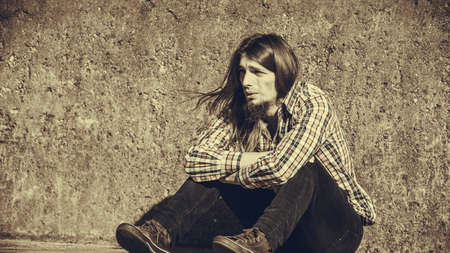 Man bearded long hair sitting sad alone by grunge wall outdoor. Unemployment depression or sadness concept.の写真素材