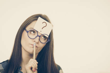 Woman confused thinking seeks a solution, paper card with question mark on her head. Doubtful young female in glasses studio shot toned imageの写真素材
