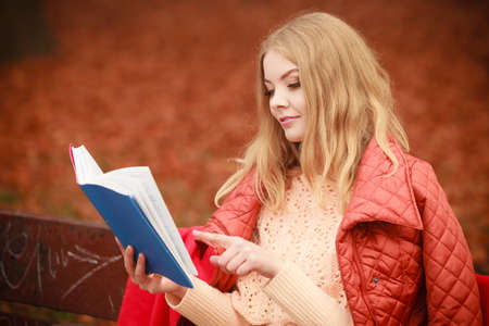 Nature outdoor relax leisure literature concept. Lady reading blue book. Young blonde girl in autumnal forest sitting on bench covered by blanket.の写真素材