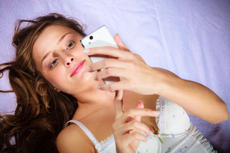 Texting and calling concept. Beauty young long haired woman lying on bad holding smartphone and texting.の写真素材