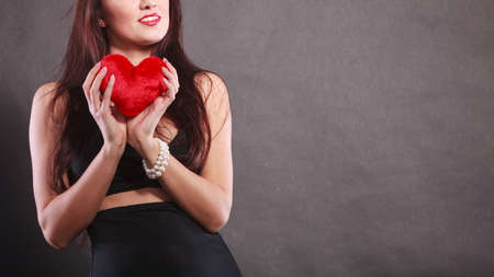 Woman brunette long hair girl wearing black sensual dress holding gift in form of red heart love symbol studio shot on dark. Valentines day happiness conceptの写真素材