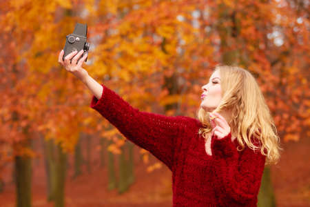 Pretty woman in fall forest park taking selfie self photo with old vintage camera. Gorgeous young girl passionate photographer blowing kiss. Autumn winter photography.の写真素材