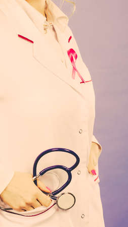 Women fight for health. Breast cancer tumor concept. Pink ribbon on white medical apron uniform and blue stethoscope in pocket.の写真素材