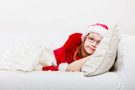 Girl family time during christmas. Cute little lady crawling on sofa. Small woman lying on the couch. Resting time wearing red santa claus hat and shoes.の写真素材