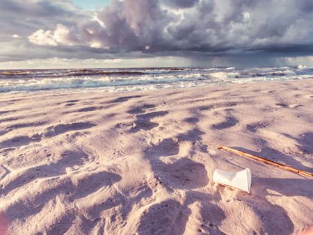 Polluted beach shore. Plastic disposable cup abandoned on sand, beautiful golden sunset in the background. Earth ecologyの写真素材