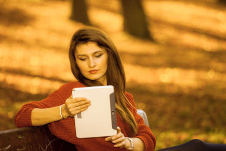 Technology and education. Woman relaxing in autumnal park sitting on bench with tablet. Pretty...