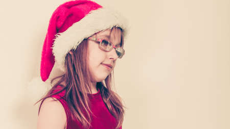 Christmas family time concept. Little girl looking like santas elf. Beautiful young lady wearing santa claus hat with white pompon. Small woman has red dress with bow and wears glasses.の写真素材