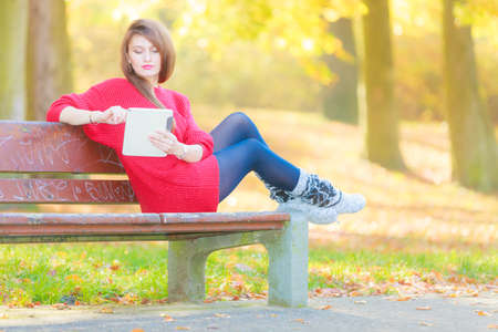 Technology and education. Woman relaxing in autumnal park sitting on bench with tablet. Pretty fashionable female student learn on fresh air.の写真素材