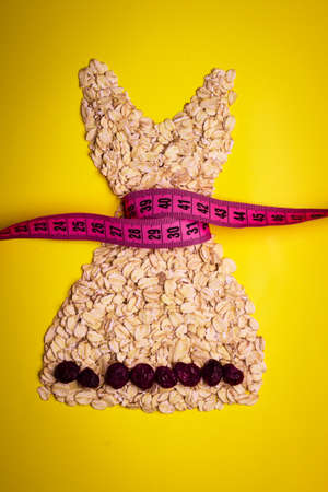 Dieting healthy eating slim down concept. Female dress shape made from oatmeal dried fruit with measuring tape around thin waist on yellowの写真素材