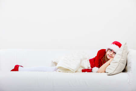 Girl family time during christmas. Cute little lady crawling on sofa. Small woman lying on the couch. Resting time wearing red santa claus hat and shoes.の写真素材