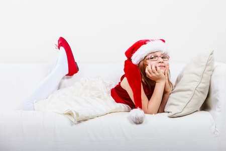 Girl family time during christmas. Cute little lady crawling on sofa. Small woman lying on the couch. Resting time wearing red santa claus hat and shoes.の写真素材