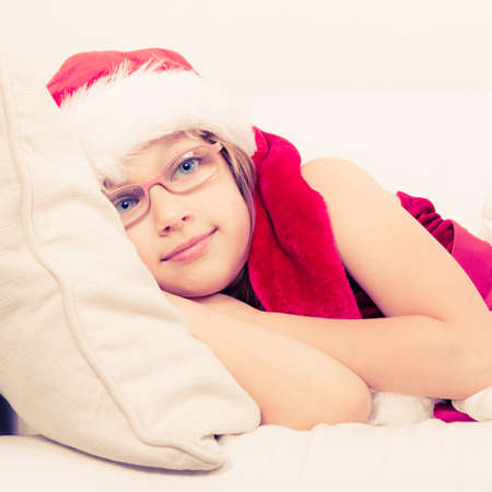 Girl family time during christmas. Cute little lady crawling on sofa. Small woman lying on the couch. Resting time wearing red santa claus hat and shoes.の写真素材