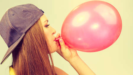 Time for party. Young trendy woman prepare birthday celebration decoration. Teenage girl in cap blowing red balloon.の写真素材
