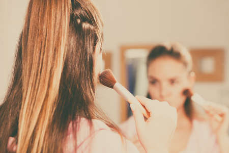 Woman face painting. Girl applying rouge or bronzing powder with brush to her skin looking in bathroom mirror. Makeup cosmetics and beauty procedures.の写真素材